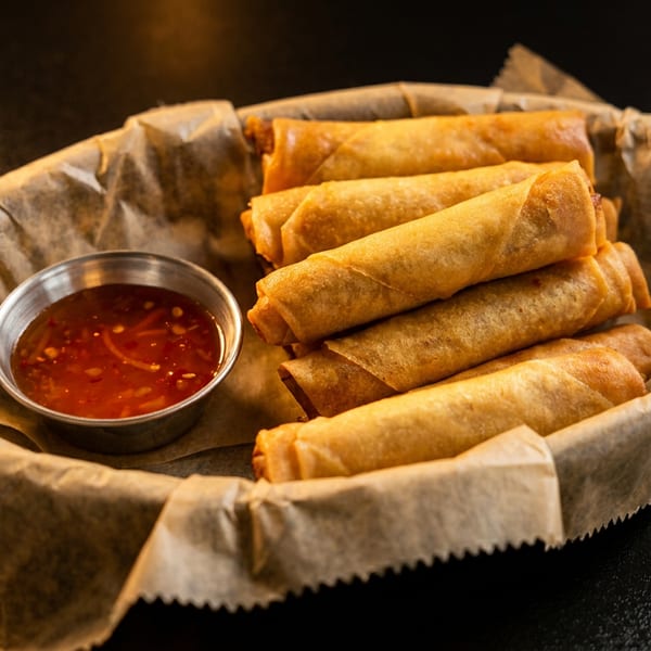 Crispy golden spring rolls served in a basket with a small cup of sweet chili dipping sauce.
