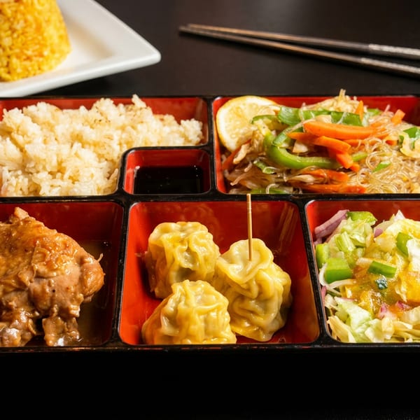 Assorted Asian bento box with chicken, shumai, rice, vegetable noodles, and fresh salad, with chopsticks.